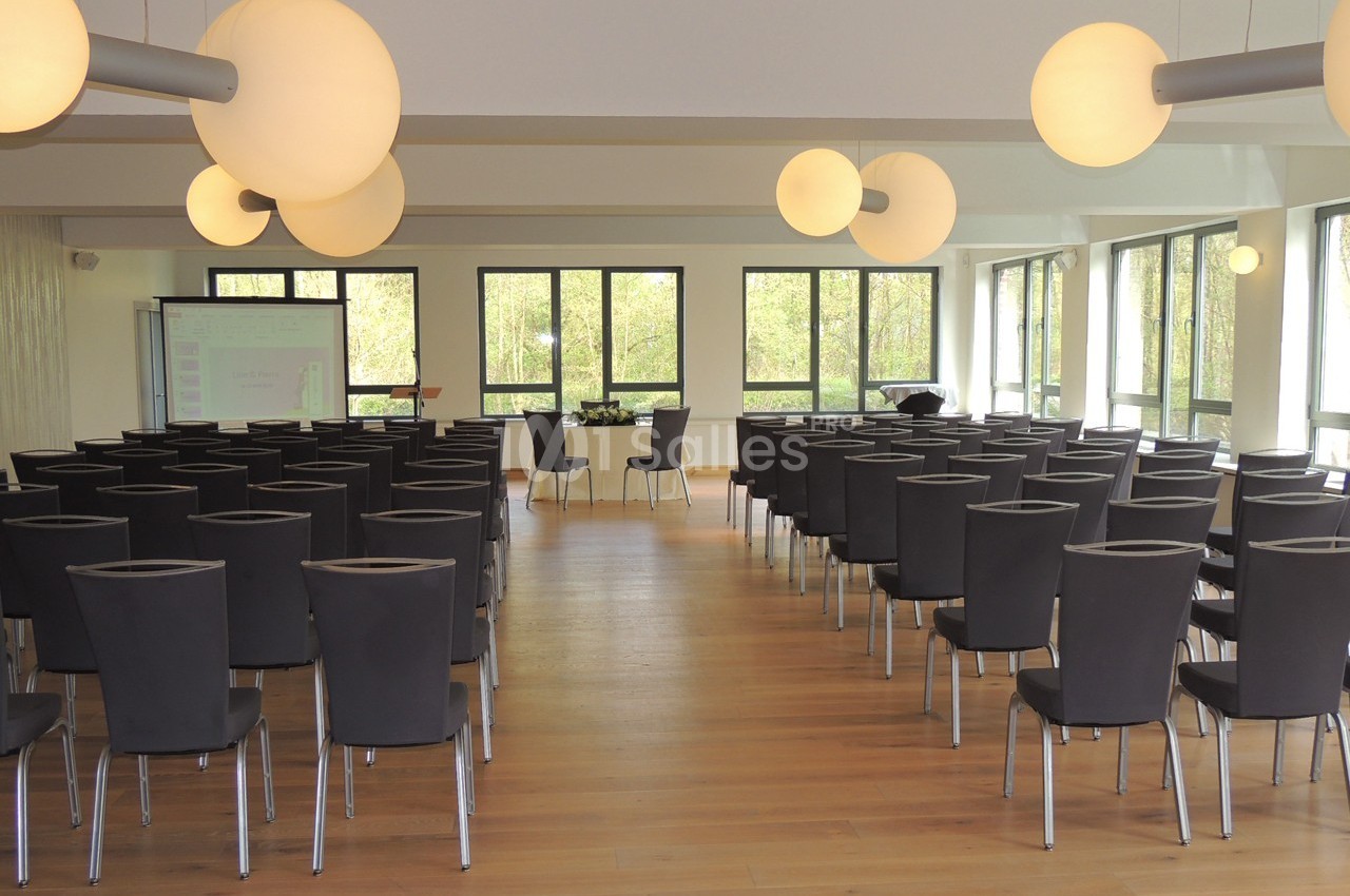Salle de conférence lumineuse avec des rangées de chaises noires, un projecteur et de grandes fenêtres donnant sur un espace…