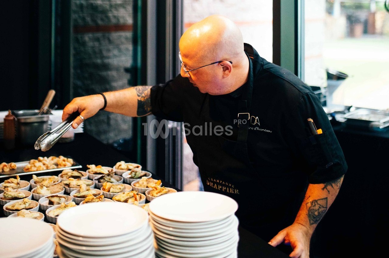 Un chef en tablier noir dispose des plats sur une table, entouré d'assiettes et d'ustensiles.