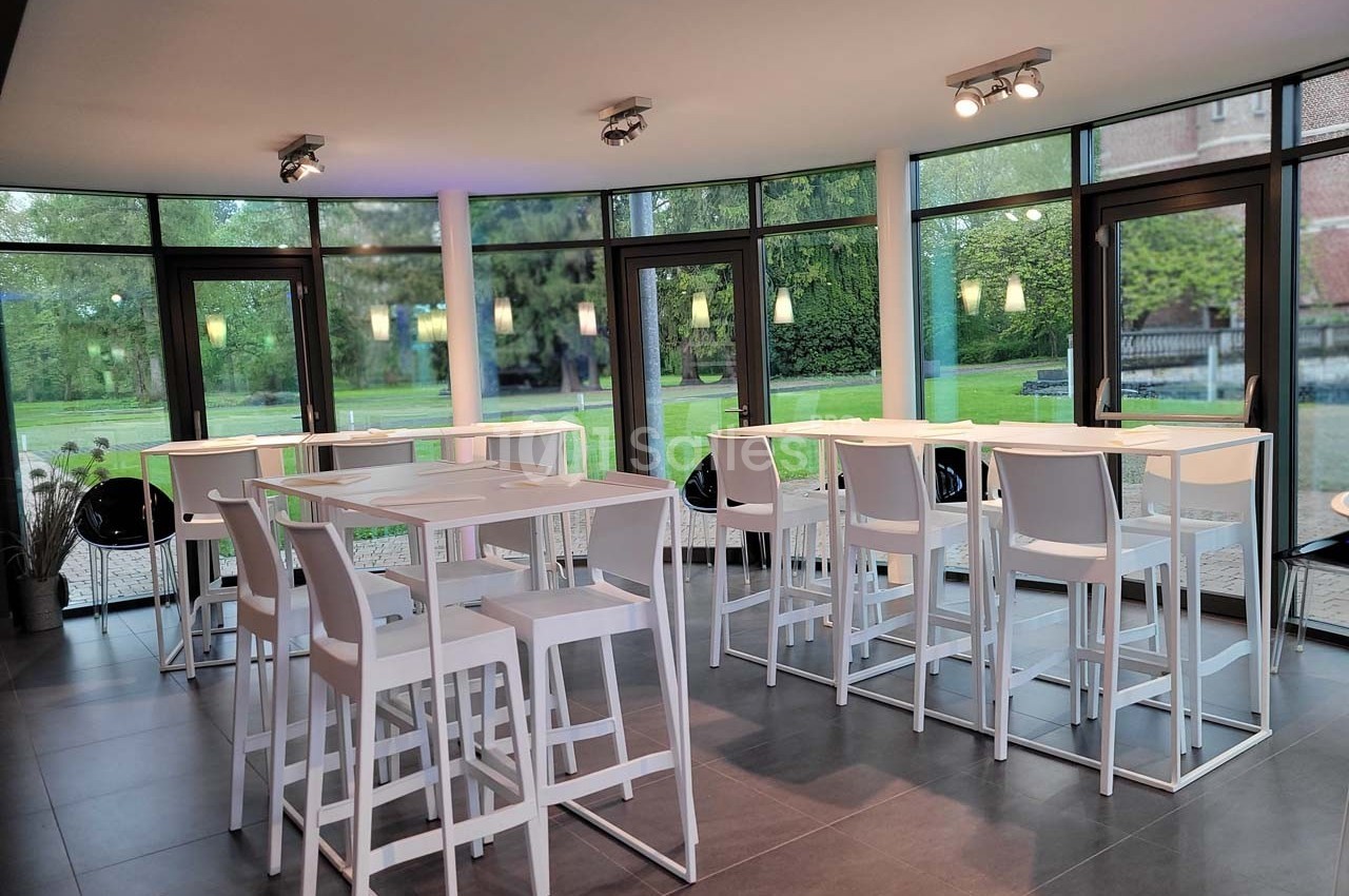 Salle lumineuse avec tables hautes blanches, chaises assorties et grandes baies vitrées donnant sur un jardin verdoyant.