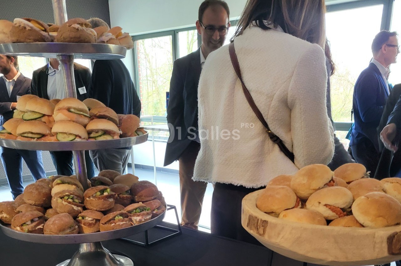 Plateaux de mini-sandwichs variés disposés sur une table lors d'un événement avec des participants en arrière-plan.