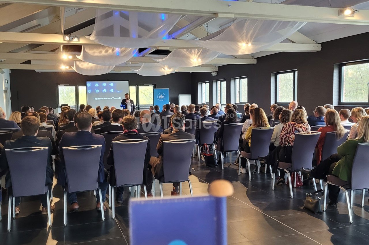 Un conférencier s'adresse à un public assis dans une salle lumineuse avec des écrans de présentation.