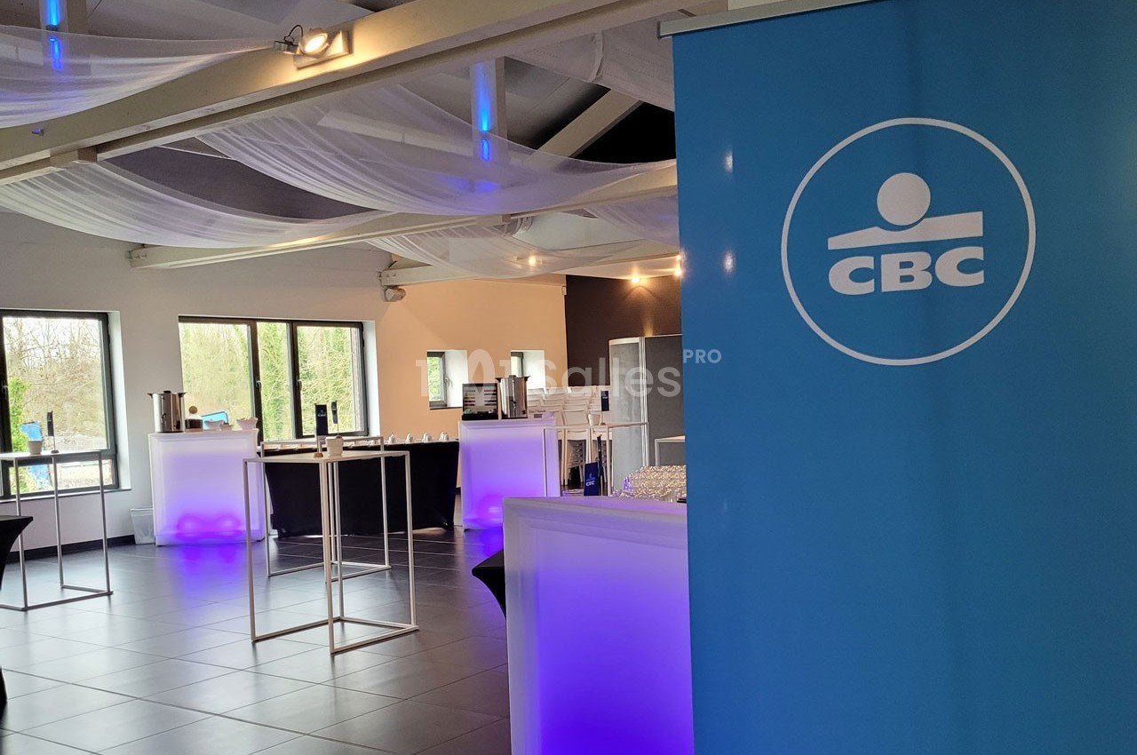 Salle lumineuse avec tables hautes, éclairage violet et un panneau bleu avec le logo ’CBC’ au premier plan.