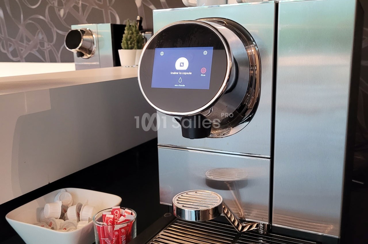 Machine à café moderne en acier inoxydable avec écran tactile, posée sur un comptoir près de dosettes et tasses.