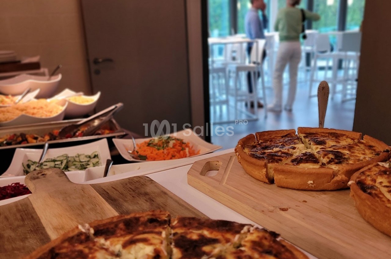 Quiches et plats variés disposés sur une table, avec deux personnes discutant en arrière-plan près de grandes fenêtres.
