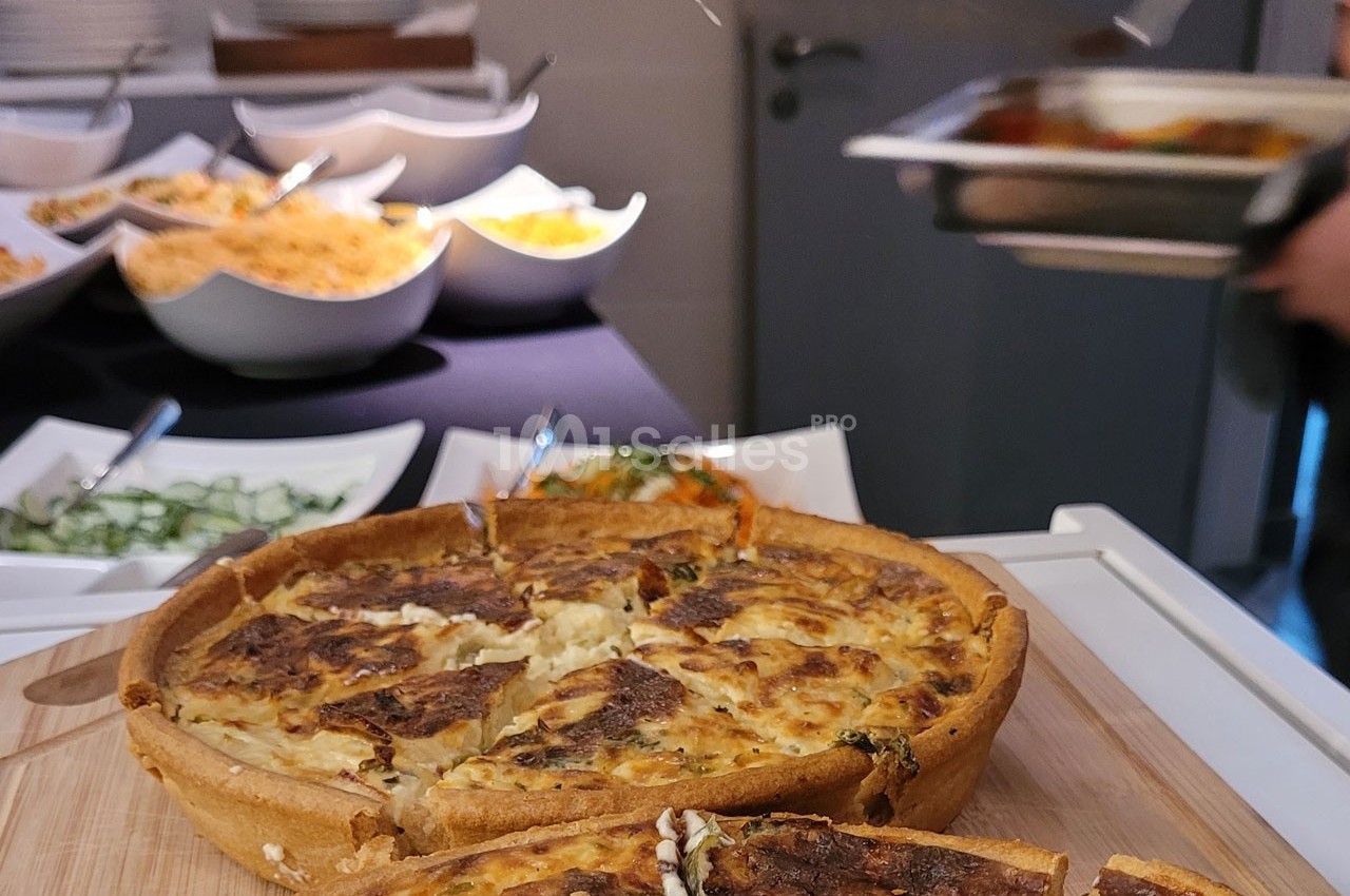 Quiche dorée coupée en parts sur une planche en bois, avec un buffet de plats variés en arrière-plan.