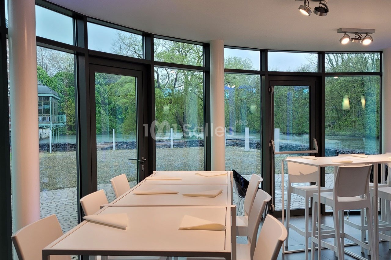 Salle lumineuse avec grandes baies vitrées donnant sur un espace extérieur arboré, tables et chaises blanches disposées.
