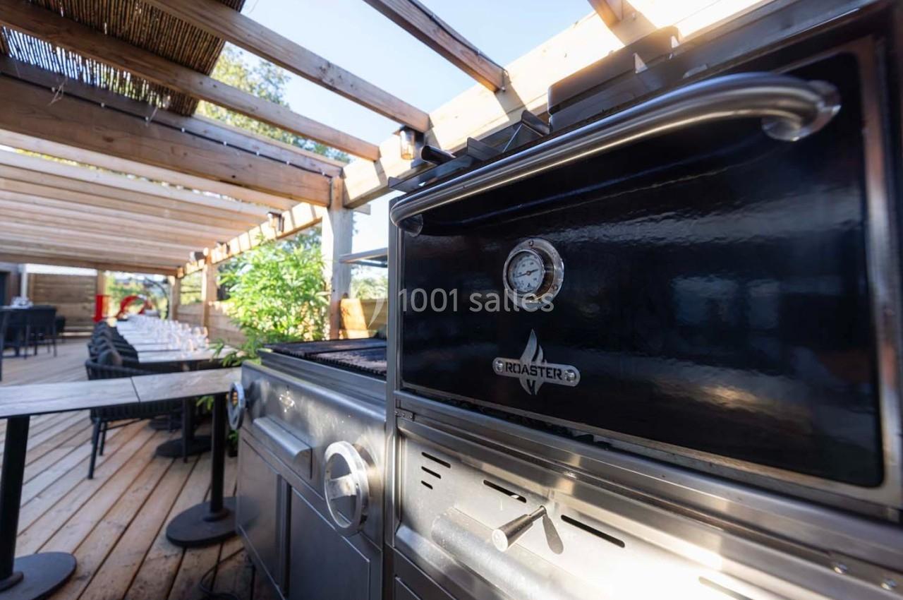 Barbecue noir sous une pergola en bois, entouré de tables et de chaises sur une terrasse ensoleillée.