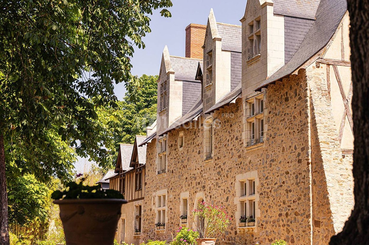 Façade en pierre d'une maison traditionnelle avec fenêtres à lucarnes, entourée d'arbres et de végétation.