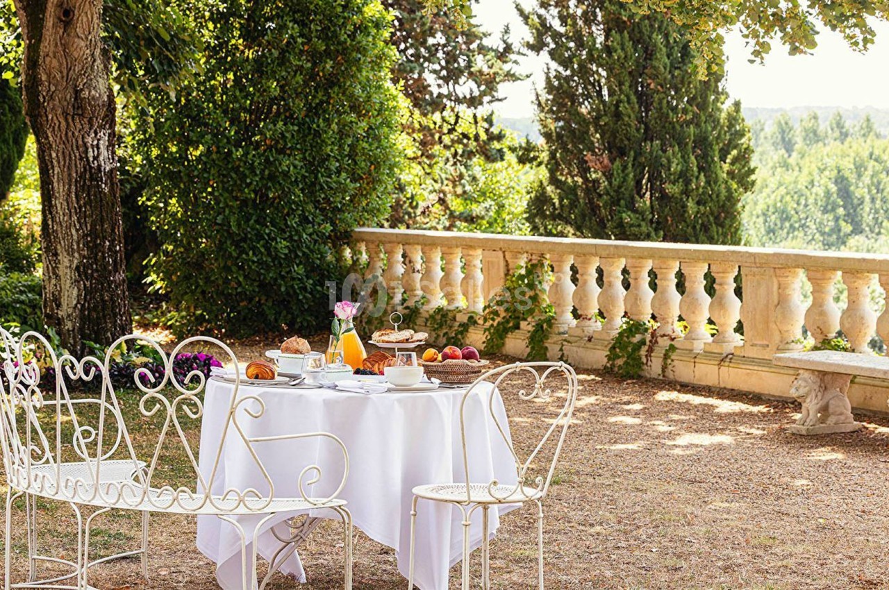 Table de jardin dressée avec petit-déjeuner, entourée de chaises en fer forgé, sous des arbres près d'une balustrade.