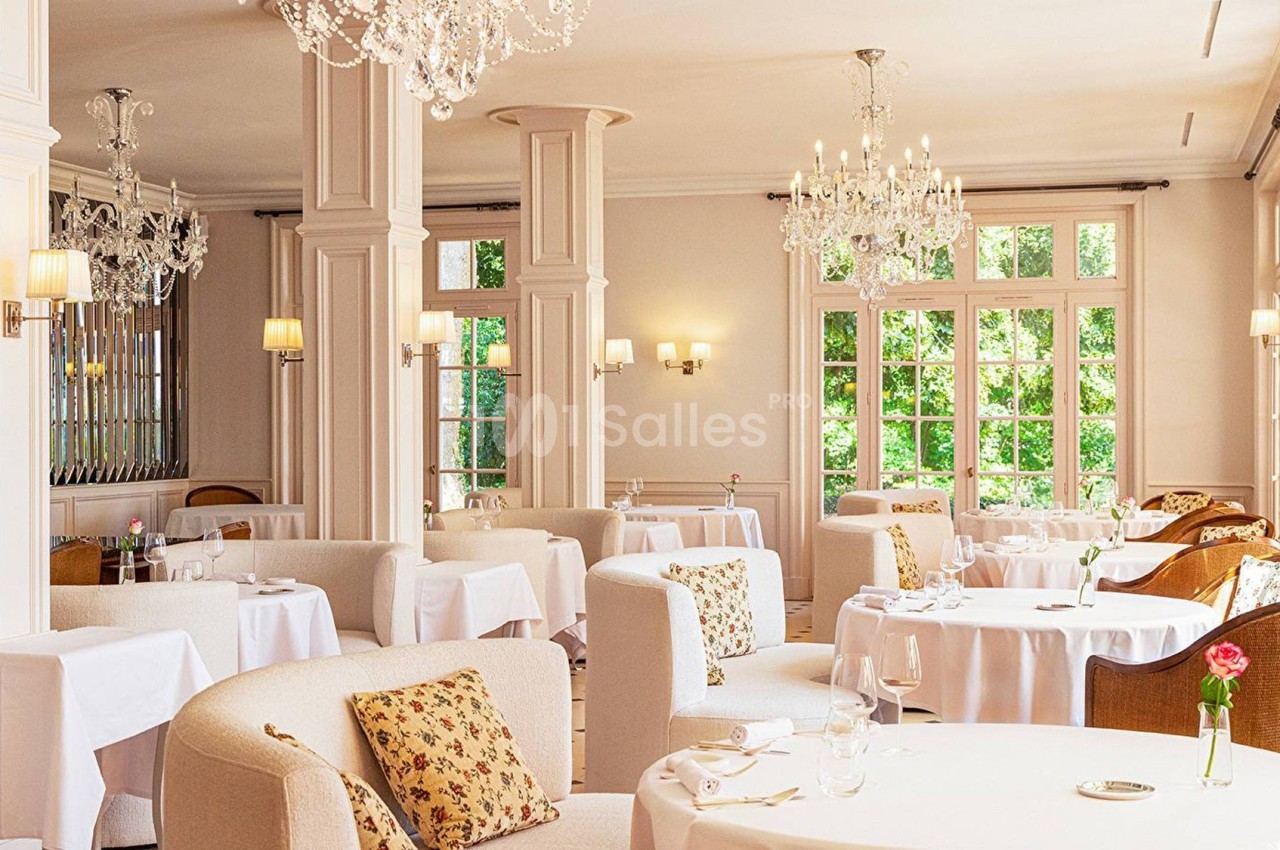 Salle de restaurant lumineuse avec tables dressées, fauteuils confortables et lustres en cristal.