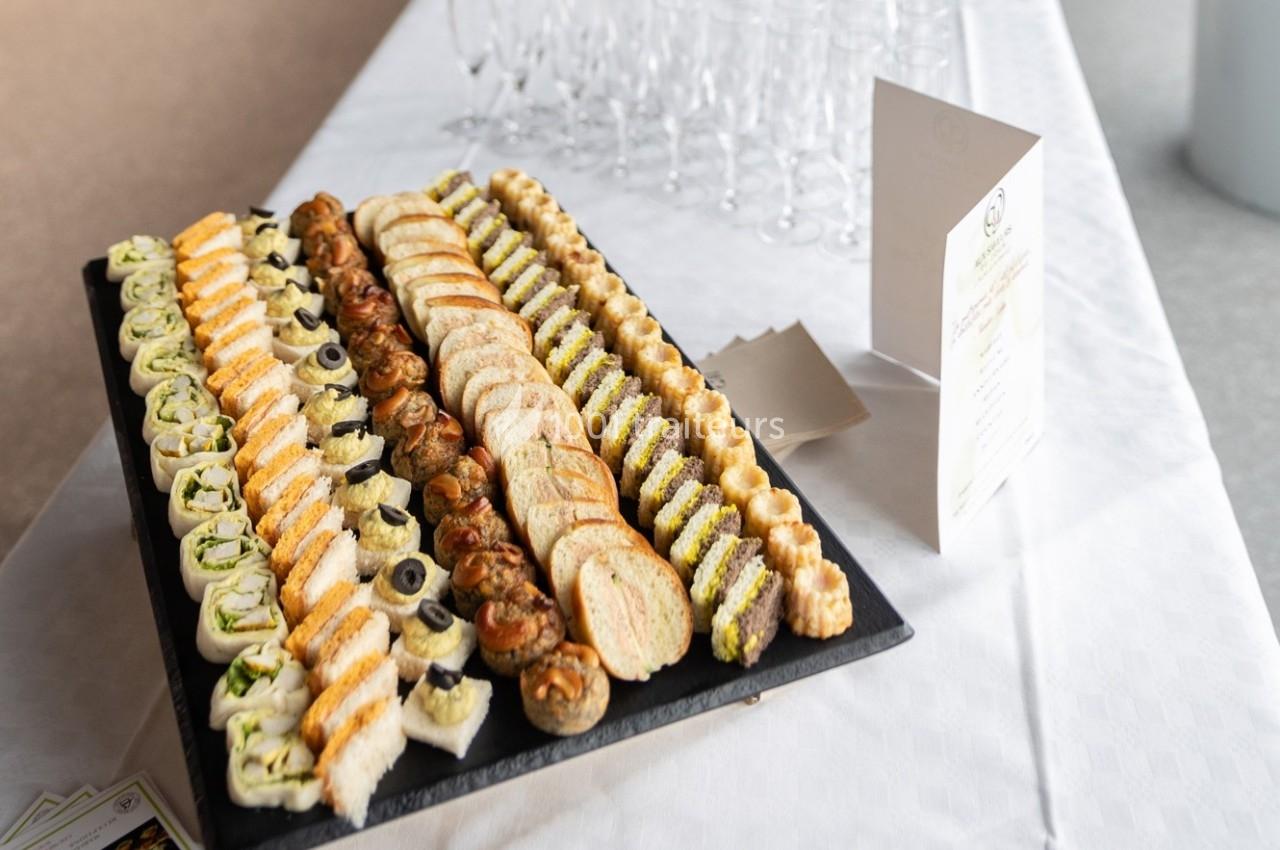 Plateau de canapés variés disposés en rangées sur une table blanche, avec des verres et un menu en arrière-plan.