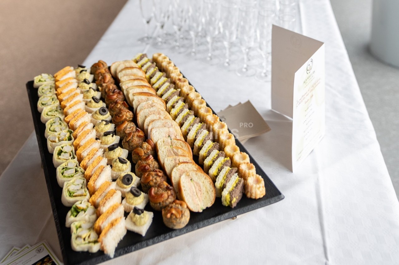 Plateau de canapés variés disposés en rangées sur une table blanche, avec des verres et un menu en arrière-plan.