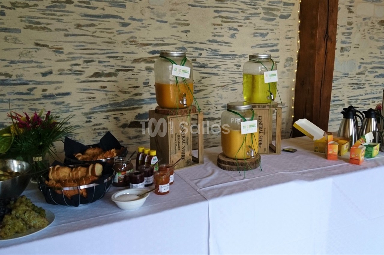 Buffet avec jus en distributeurs, viennoiseries, fruits, confitures et boissons chaudes sur une table blanche.
