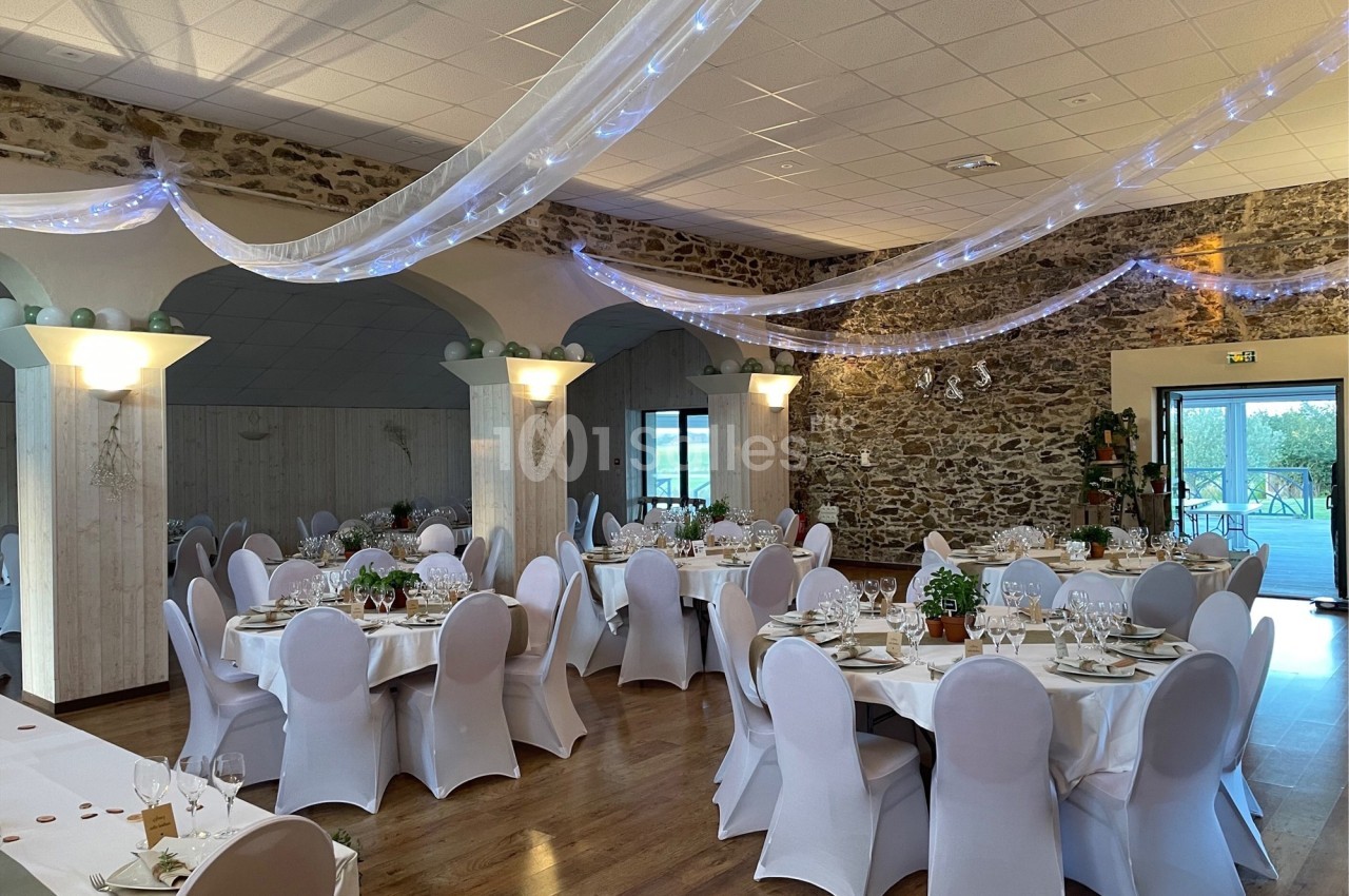 Salle de réception décorée avec des tables rondes dressées, des chaises blanches et des guirlandes lumineuses suspendues.