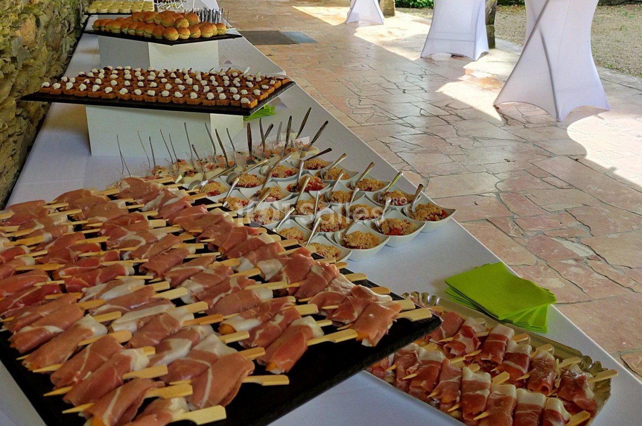 Buffet avec brochettes de jambon cru, verrines colorées et amuse-bouches disposés sur des tables en extérieur.