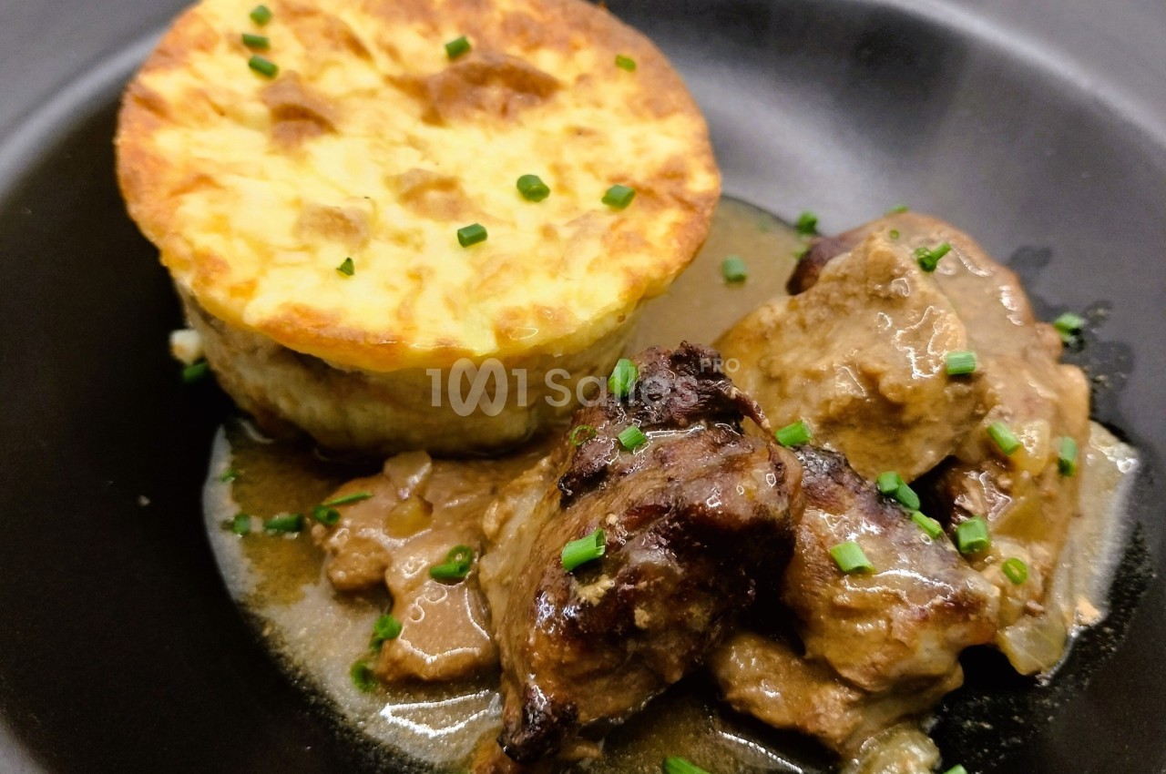 Plat composé d'un gratin doré et de morceaux de viande en sauce, servi dans une assiette noire avec ciboulette.