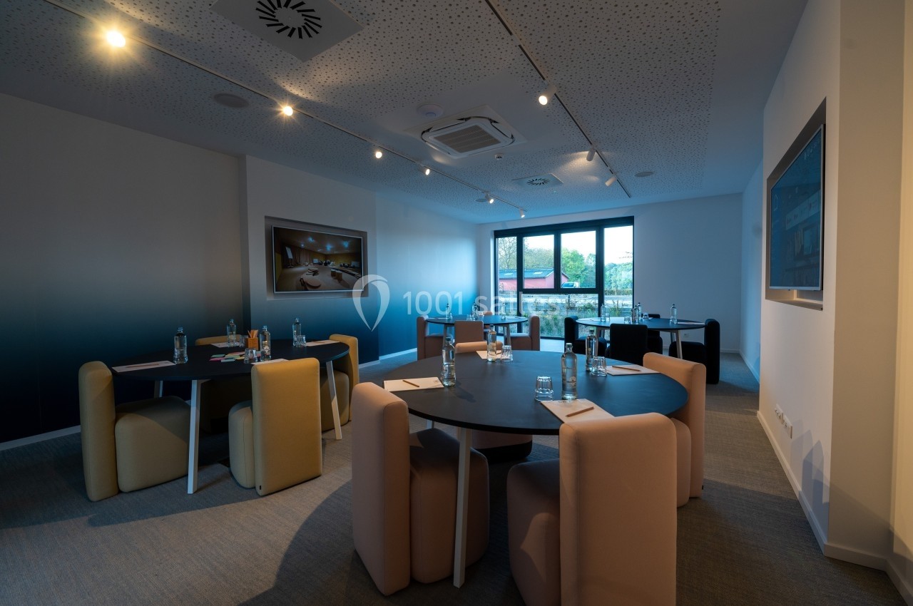Salle de réunion moderne avec tables rondes, chaises rembourrées et lumière naturelle provenant d'une grande fenêtre.