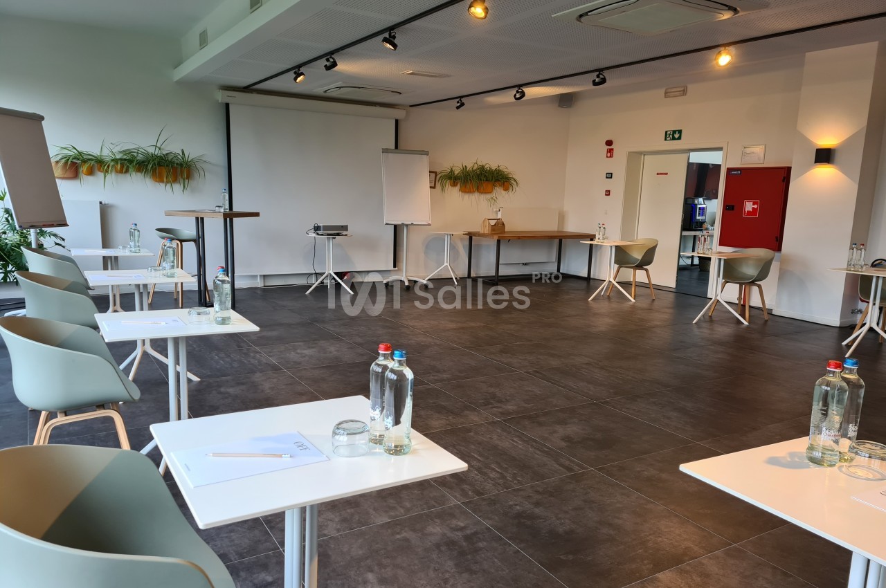 Salle de réunion lumineuse avec tables espacées, chaises modernes, tableau blanc et bouteilles d'eau sur les tables.