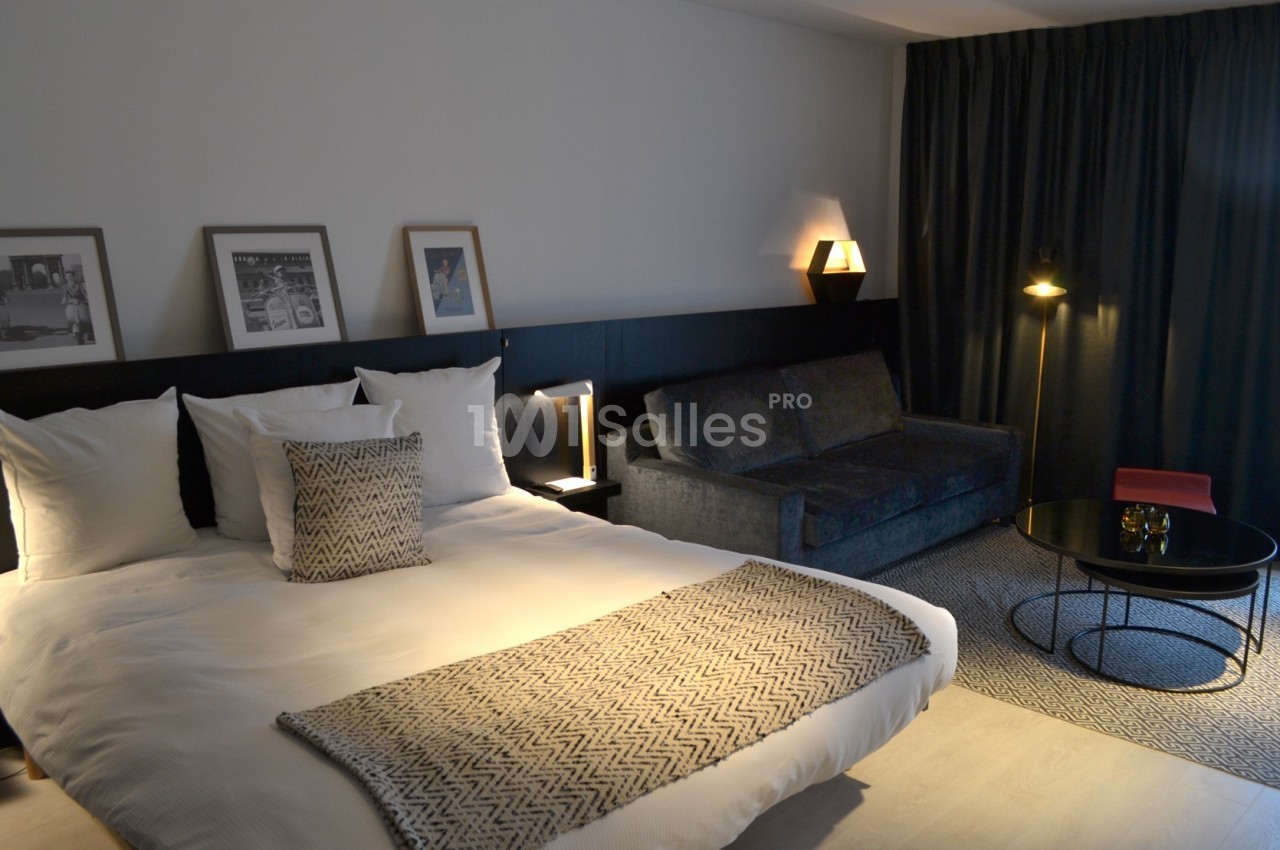 Chambre moderne avec lit double, canapé, table basse et éclairage tamisé, décorée de cadres et rideaux sombres.