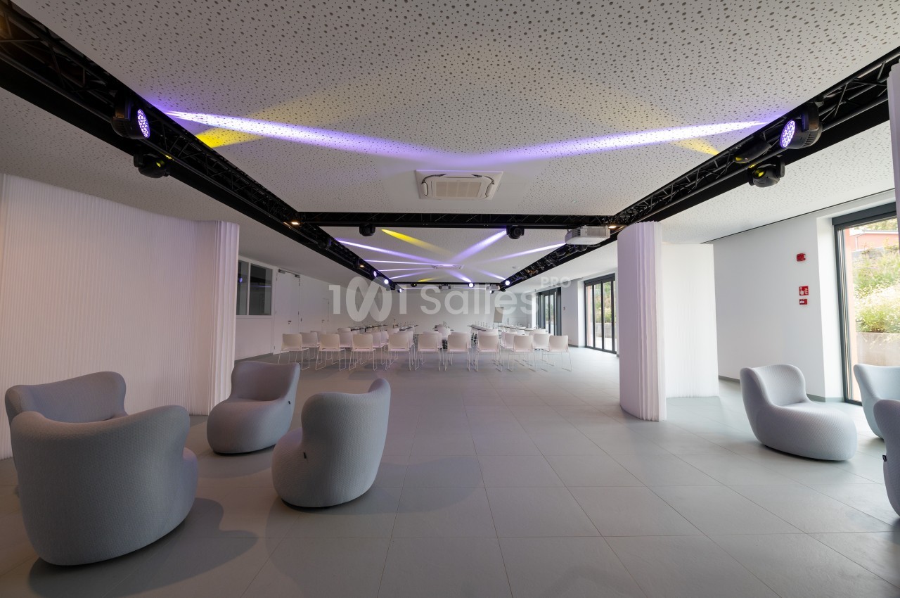 Salle lumineuse avec fauteuils modernes, tables et chaises alignées, éclairage au plafond et grandes baies vitrées.