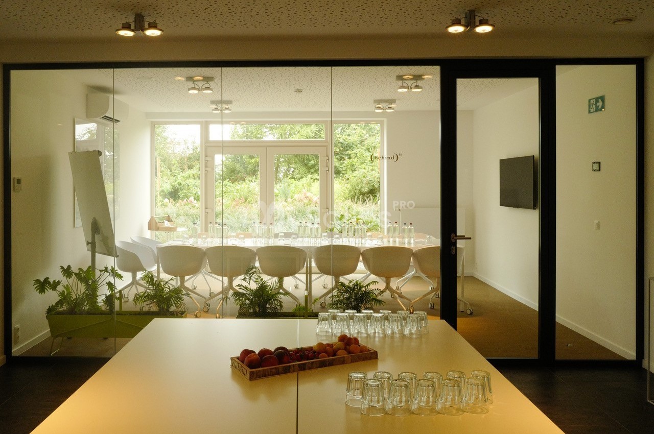 Salle de réunion lumineuse avec une grande table entourée de chaises blanches, vue sur un jardin à travers des baies vitrées.