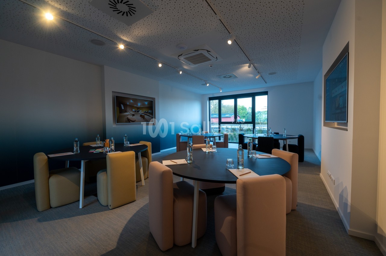 Salle de réunion moderne avec tables rondes, chaises rembourrées et lumière naturelle provenant d'une grande fenêtre.