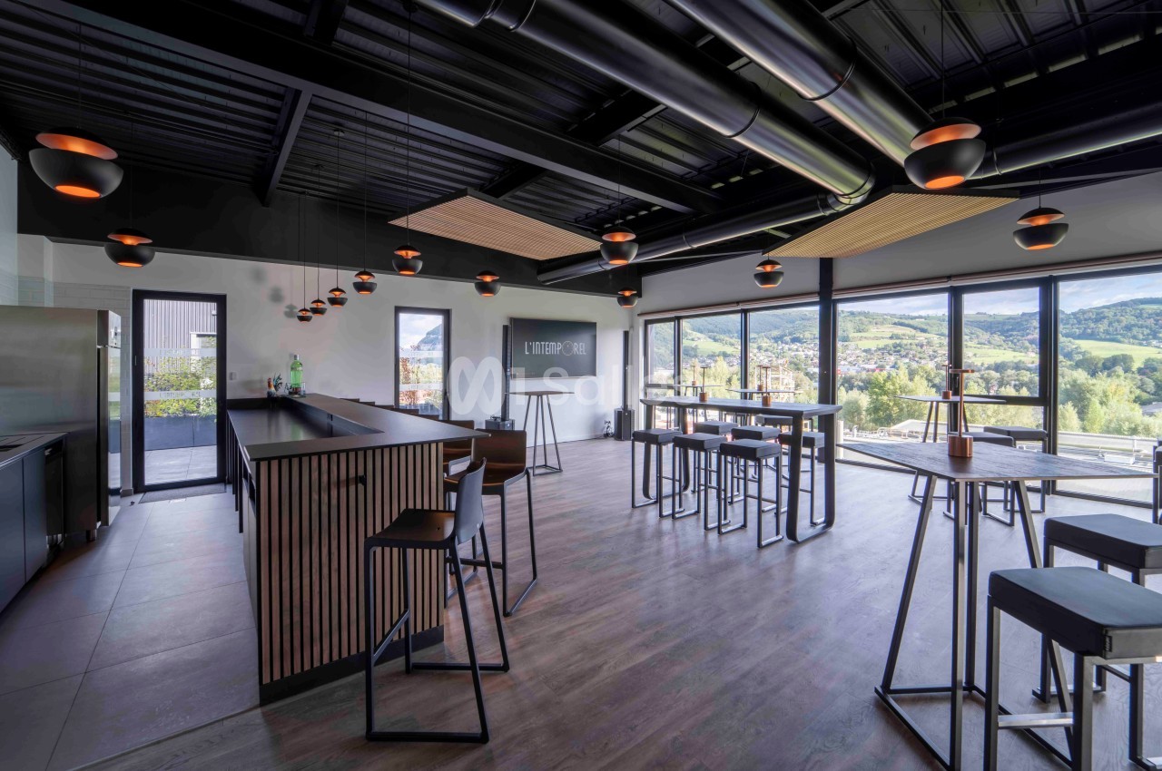 Salle moderne avec bar, tables hautes et grandes baies vitrées offrant une vue sur un paysage vallonné.