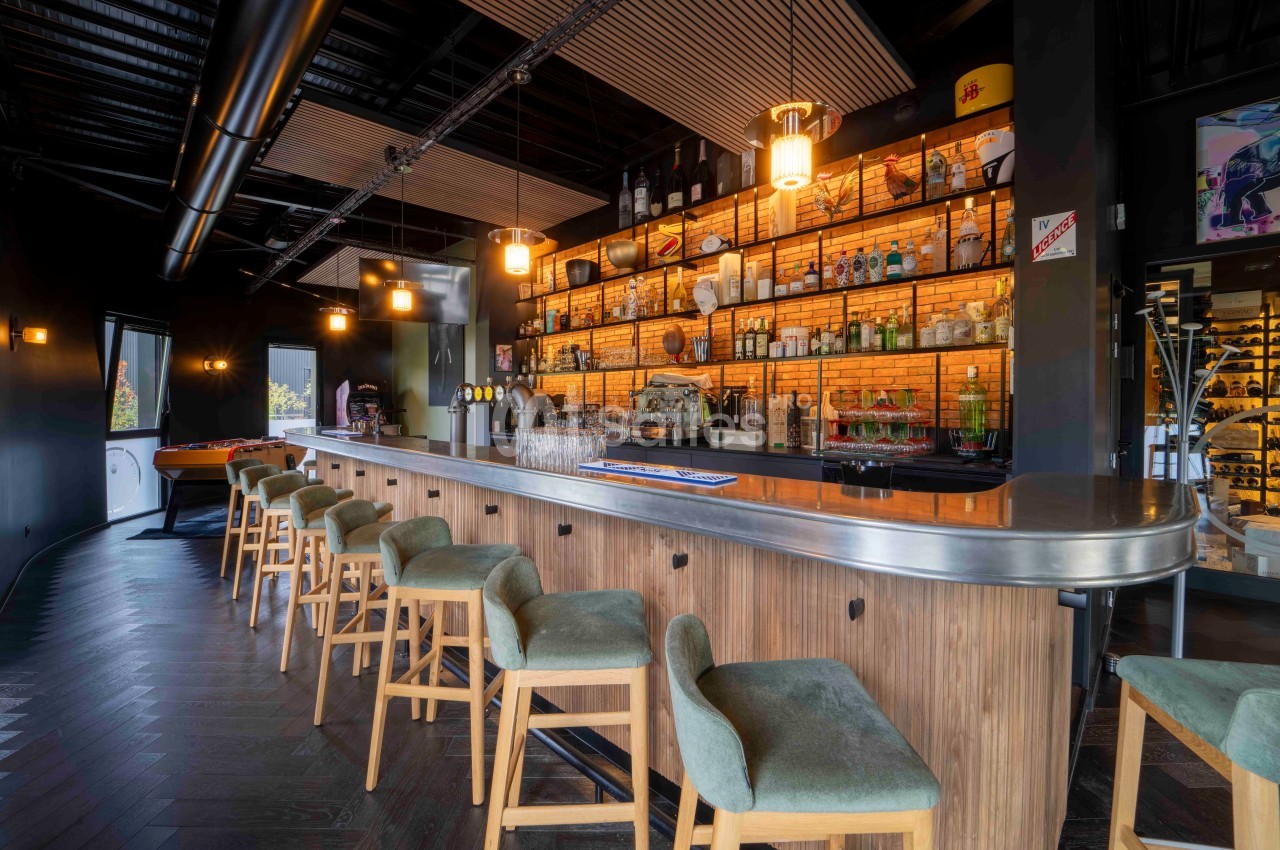 Bar moderne avec comptoir en bois, tabourets rembourrés verts, étagères éclairées et décorations murales.
