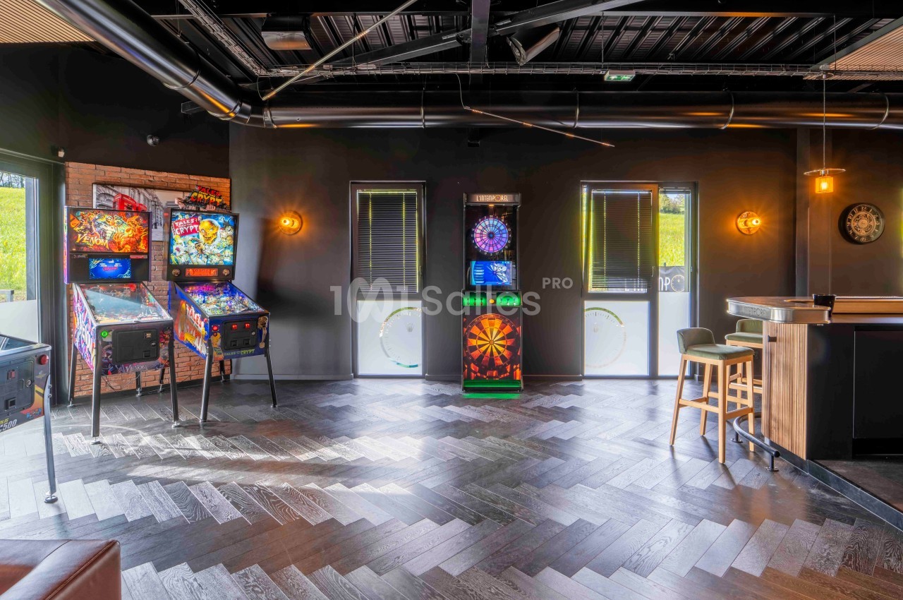 Salle de jeux avec flippers, cible de fléchettes électronique et mobilier moderne sur un sol en parquet sombre.
