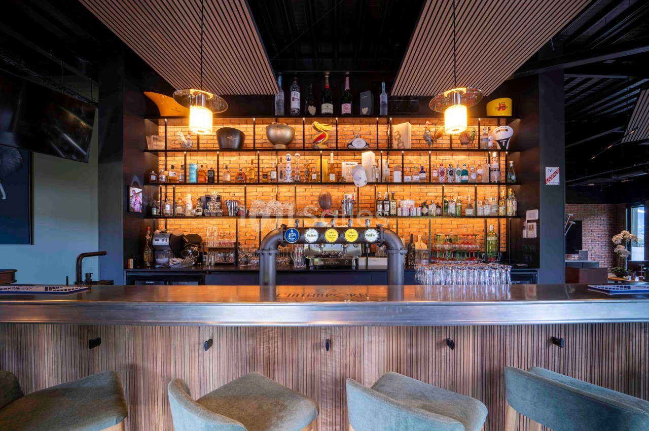 Bar moderne avec comptoir en bois, tabourets verts et étagères éclairées exposant des bouteilles et des verres.
