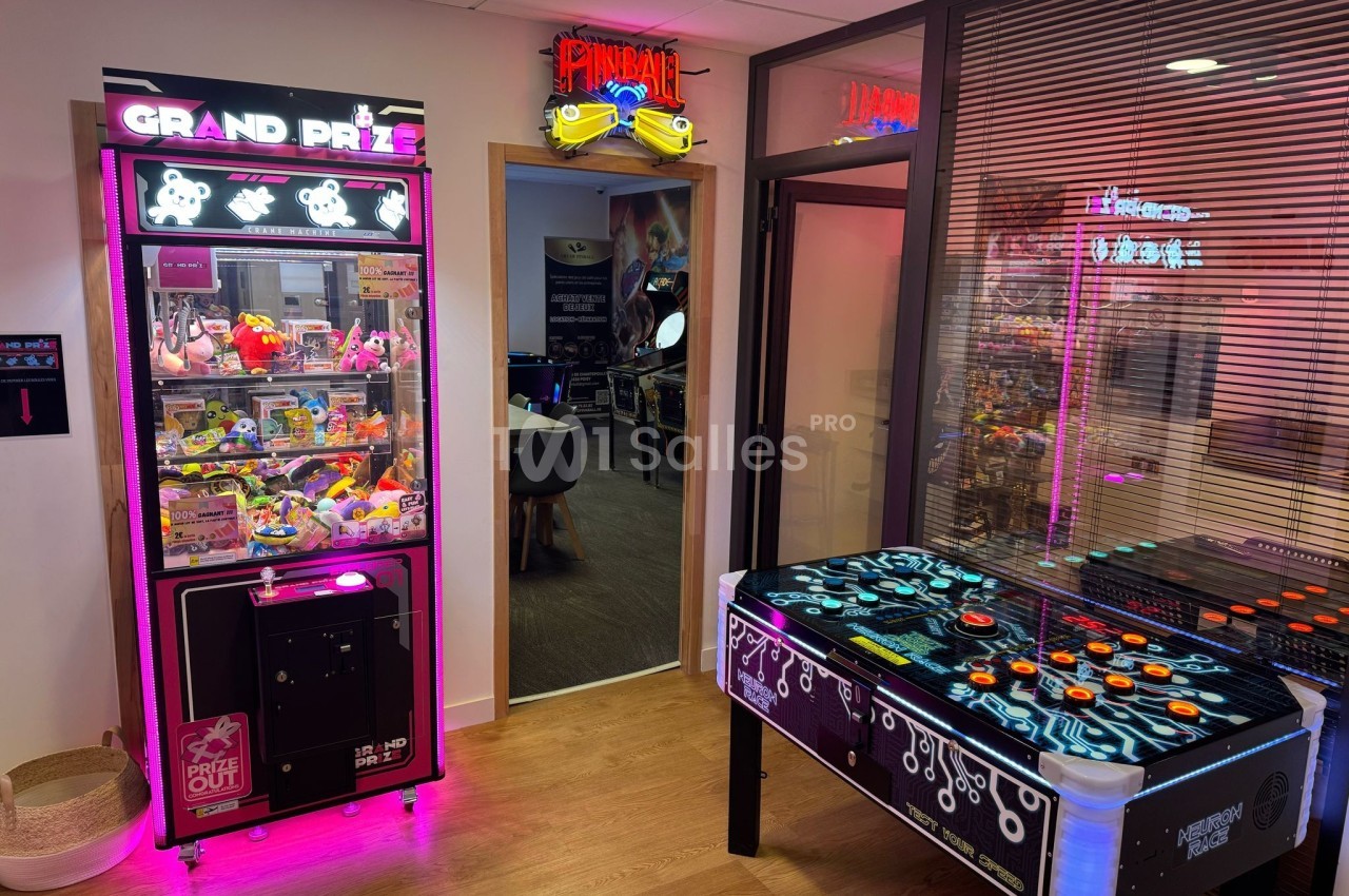 Salle avec une machine à pinces lumineuse, une table de jeu rétro et une enseigne ’Pinball’ au-dessus d'une porte.