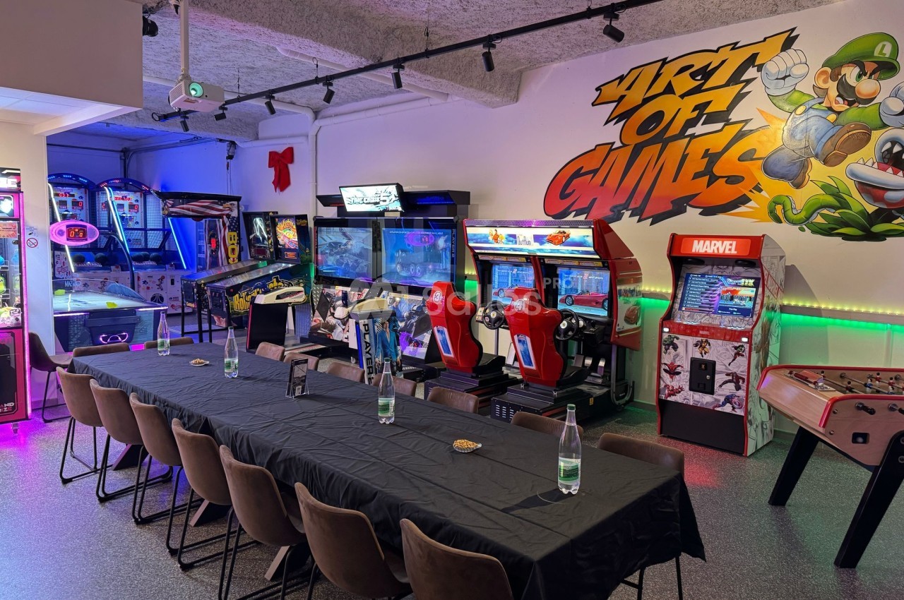 Salle d'arcade avec bornes de jeux, flippers, simulateurs de course et table dressée pour un événement.