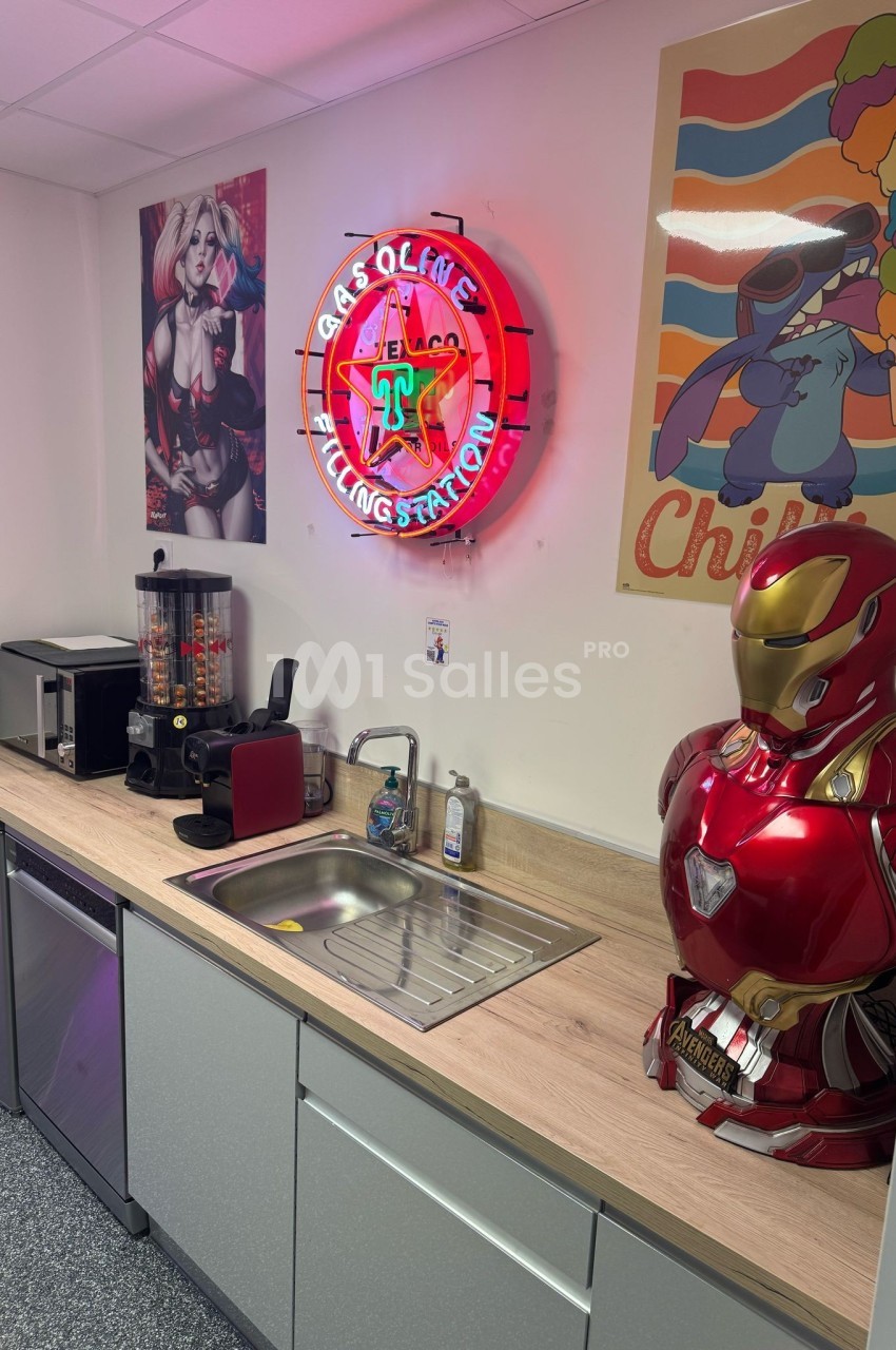 Cuisine équipée avec évier, cafetières, distributeur de bonbons, déco néon et posters, et buste d'armure rouge.