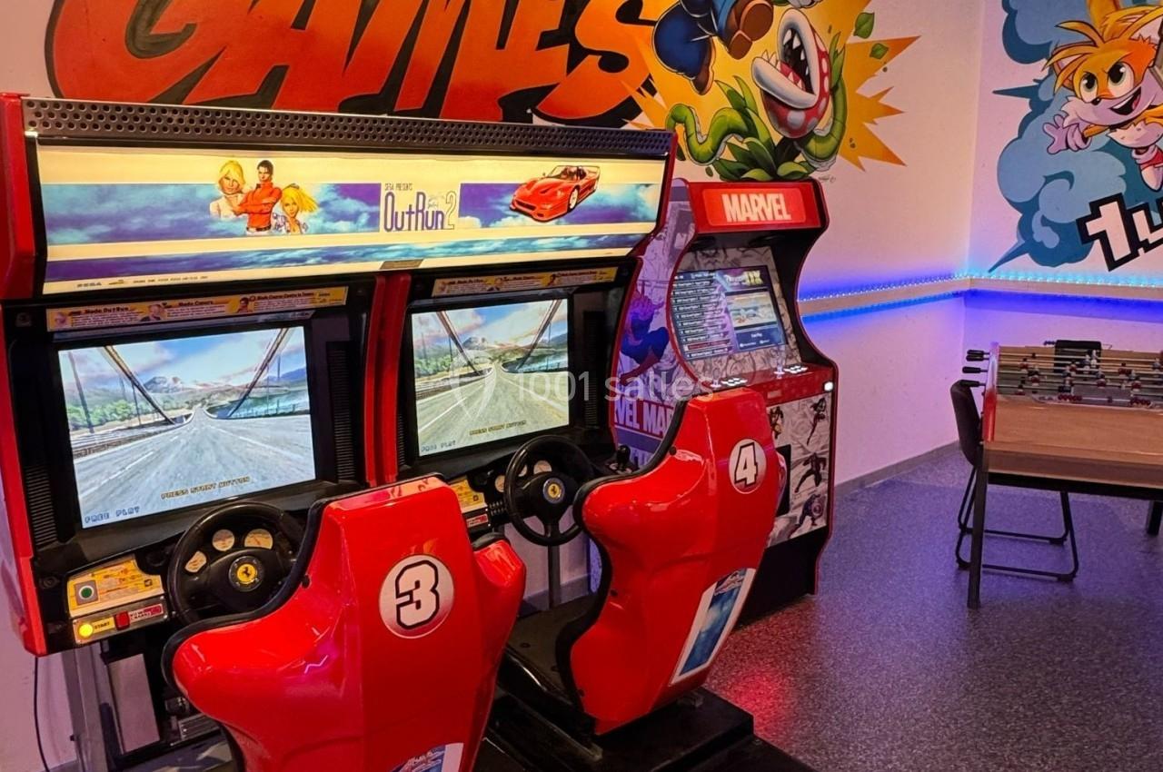 Deux bornes d'arcade rétro avec sièges rouges, un flipper Marvel et une table dans une salle décorée de fresques colorées.