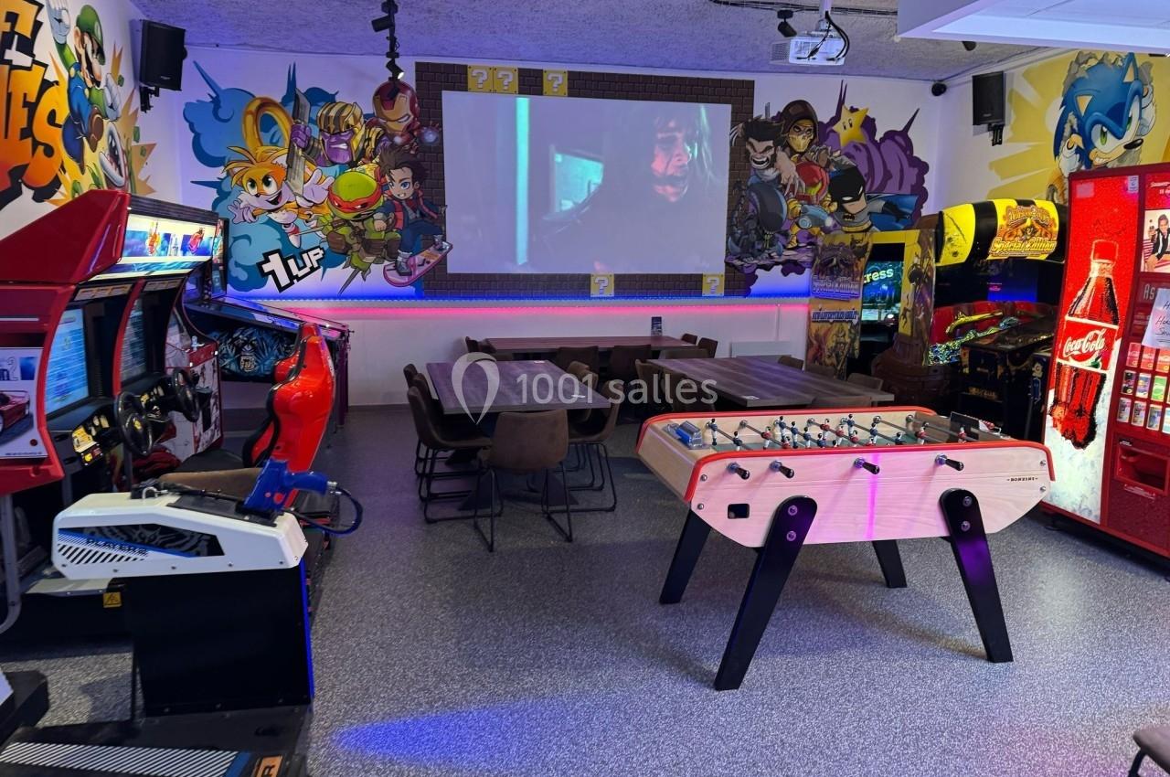 Salle de jeux avec baby-foot, bornes d'arcade, tables, écran projetant un film et décorations murales colorées.