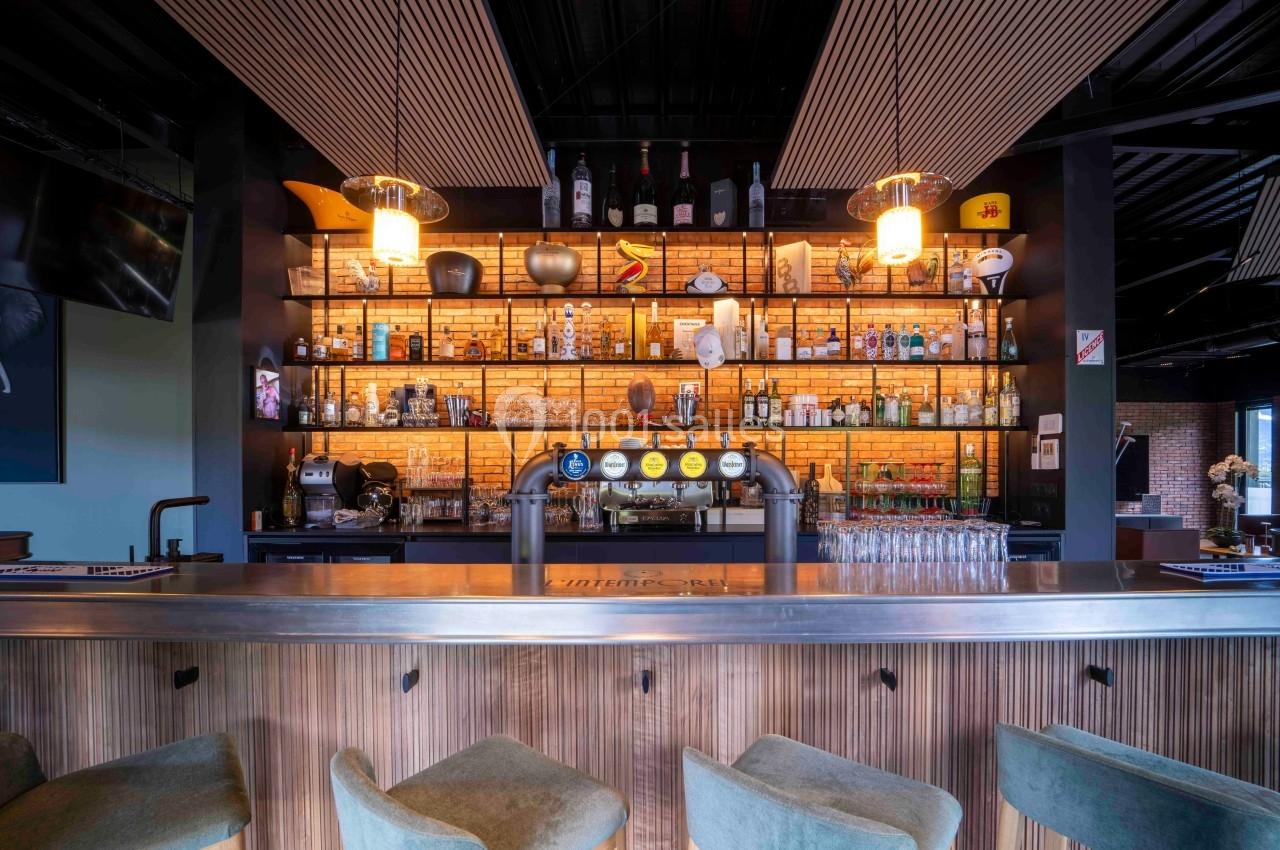 Bar moderne avec comptoir en bois, tabourets verts et étagères illuminées exposant bouteilles et verres.