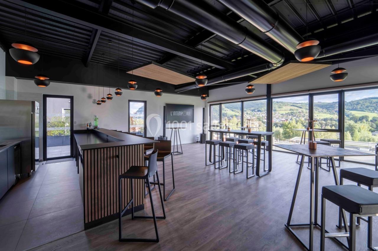 Salle moderne avec bar, tables hautes et grandes baies vitrées offrant une vue sur un paysage vallonné.