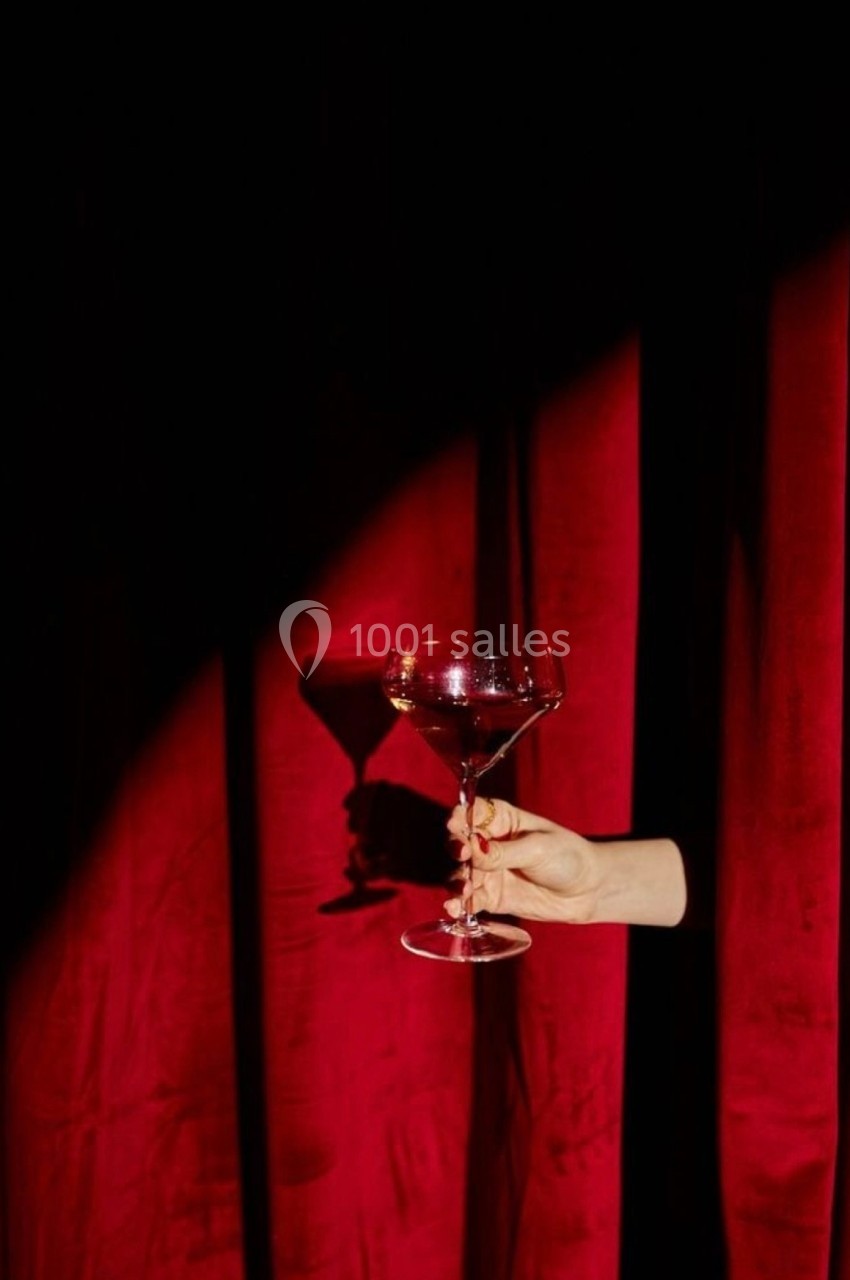 Une main tient une coupe de champagne devant un rideau rouge éclairé par une lumière tamisée.