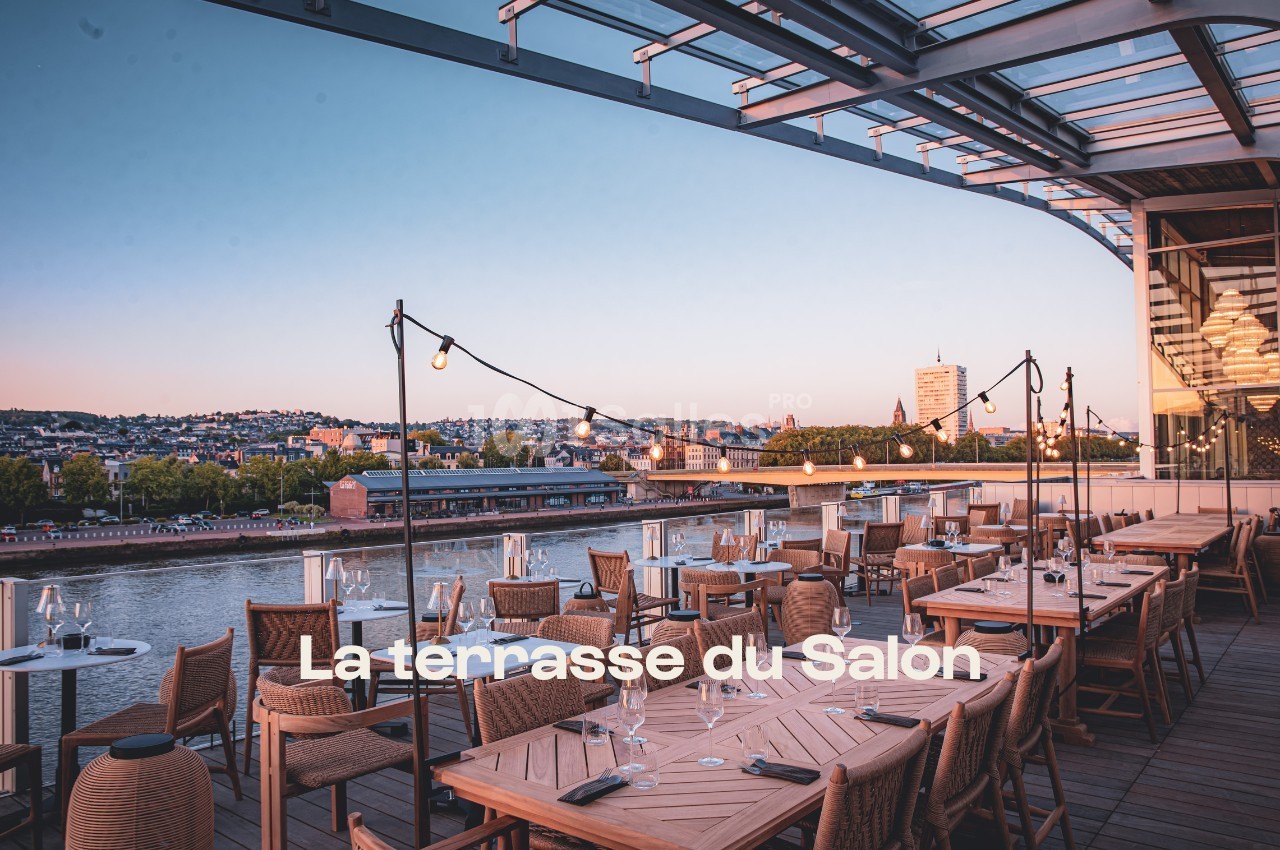 Terrasse en bois avec tables dressées, vue sur un fleuve et une ville au coucher du soleil.
