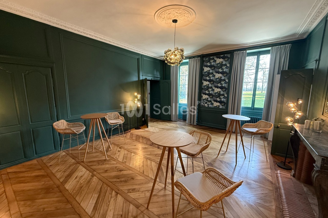 Salle lumineuse avec parquet en bois, murs verts, tables hautes et chaises en rotin, vue sur un jardin à travers les…