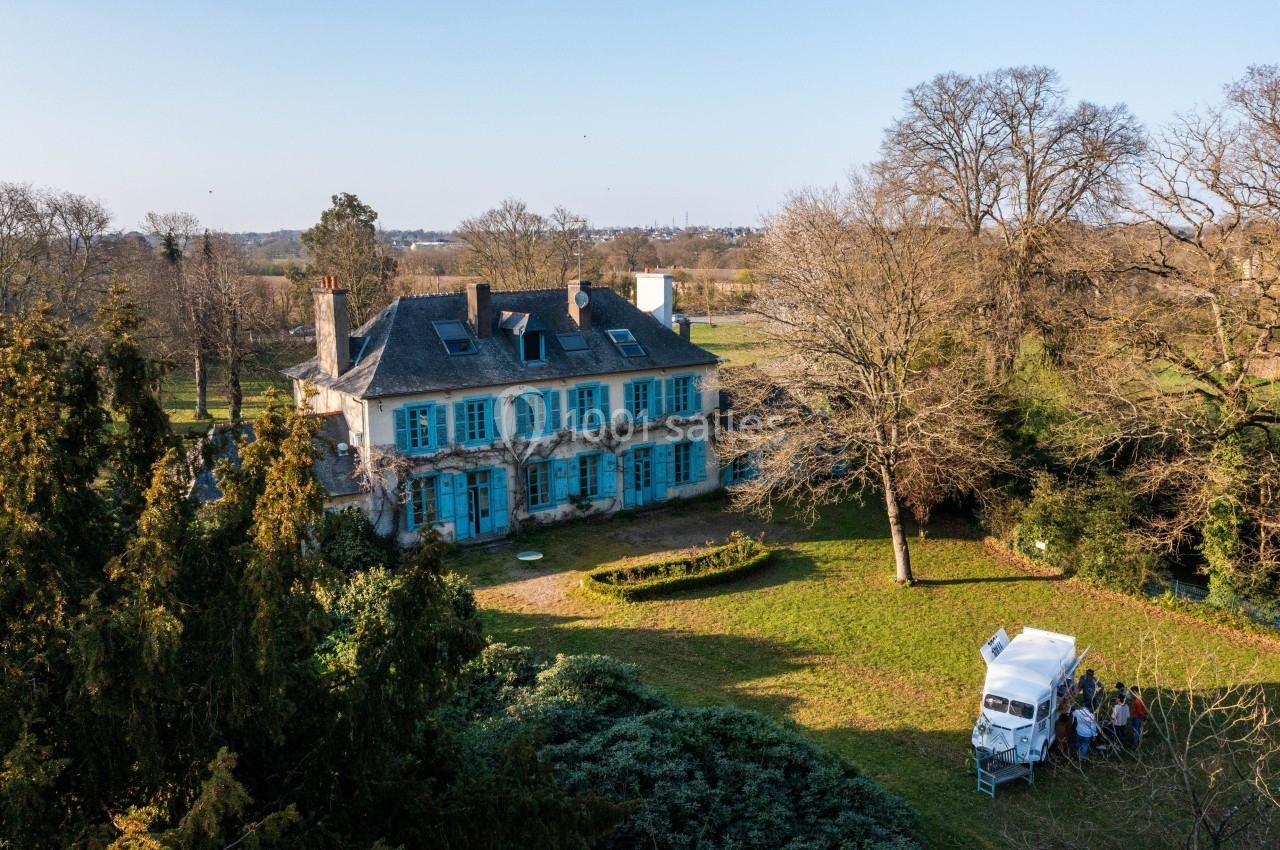 Manoir ancien avec volets bleus entouré de jardins, vue aérienne par temps clair, petit groupe près d'un véhicule blanc.