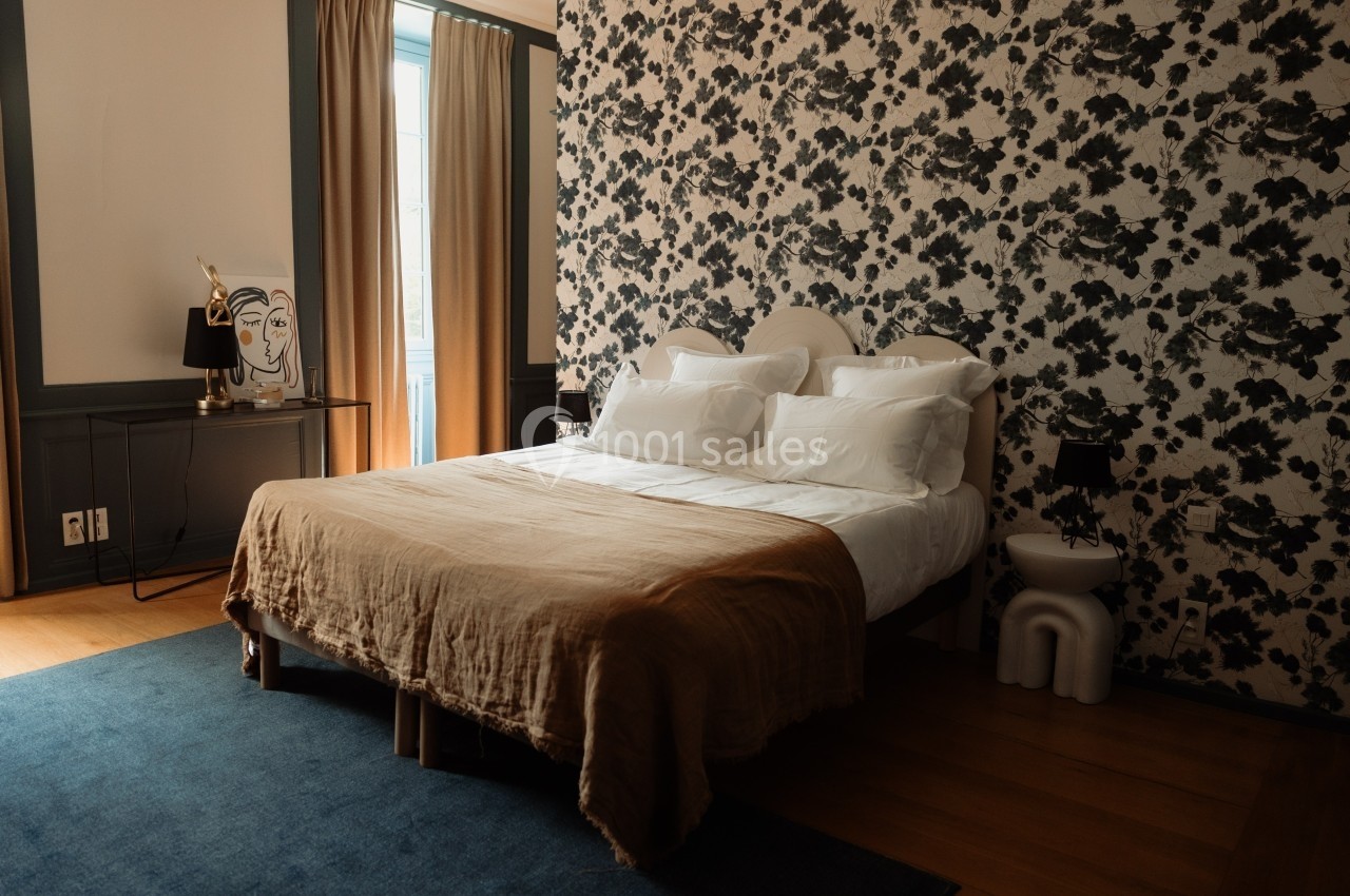 Chambre avec lit double, couvre-lit beige, papier peint floral noir et blanc, rideaux beige et lumière naturelle.