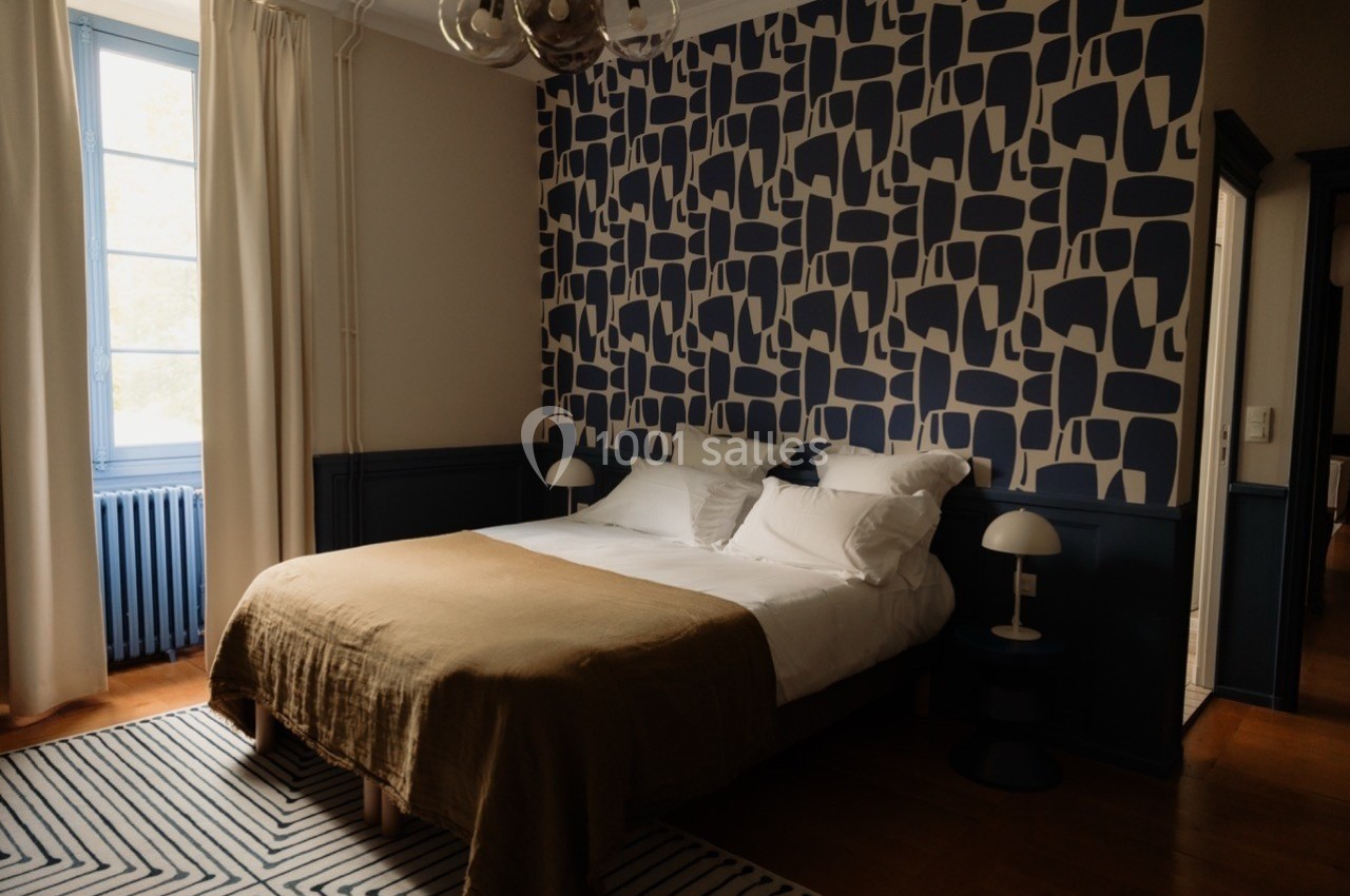 Chambre avec lit double, mur décoré de motifs géométriques bleus et blancs, lumière naturelle entrant par une fenêtre.
