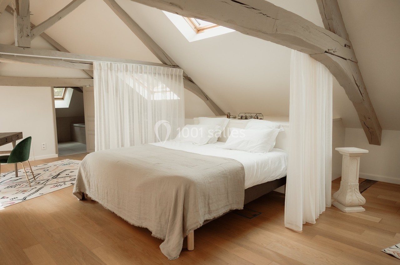 Chambre lumineuse sous combles avec lit double, poutres apparentes, parquet clair et rideaux blancs.