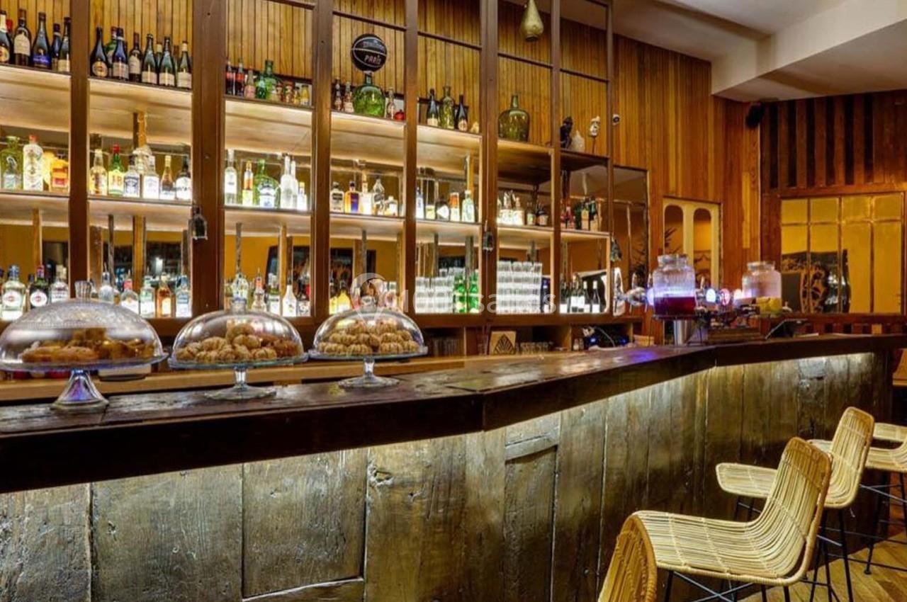Intérieur d'un bar avec comptoir en bois, étagères remplies de bouteilles et chaises en rotin.