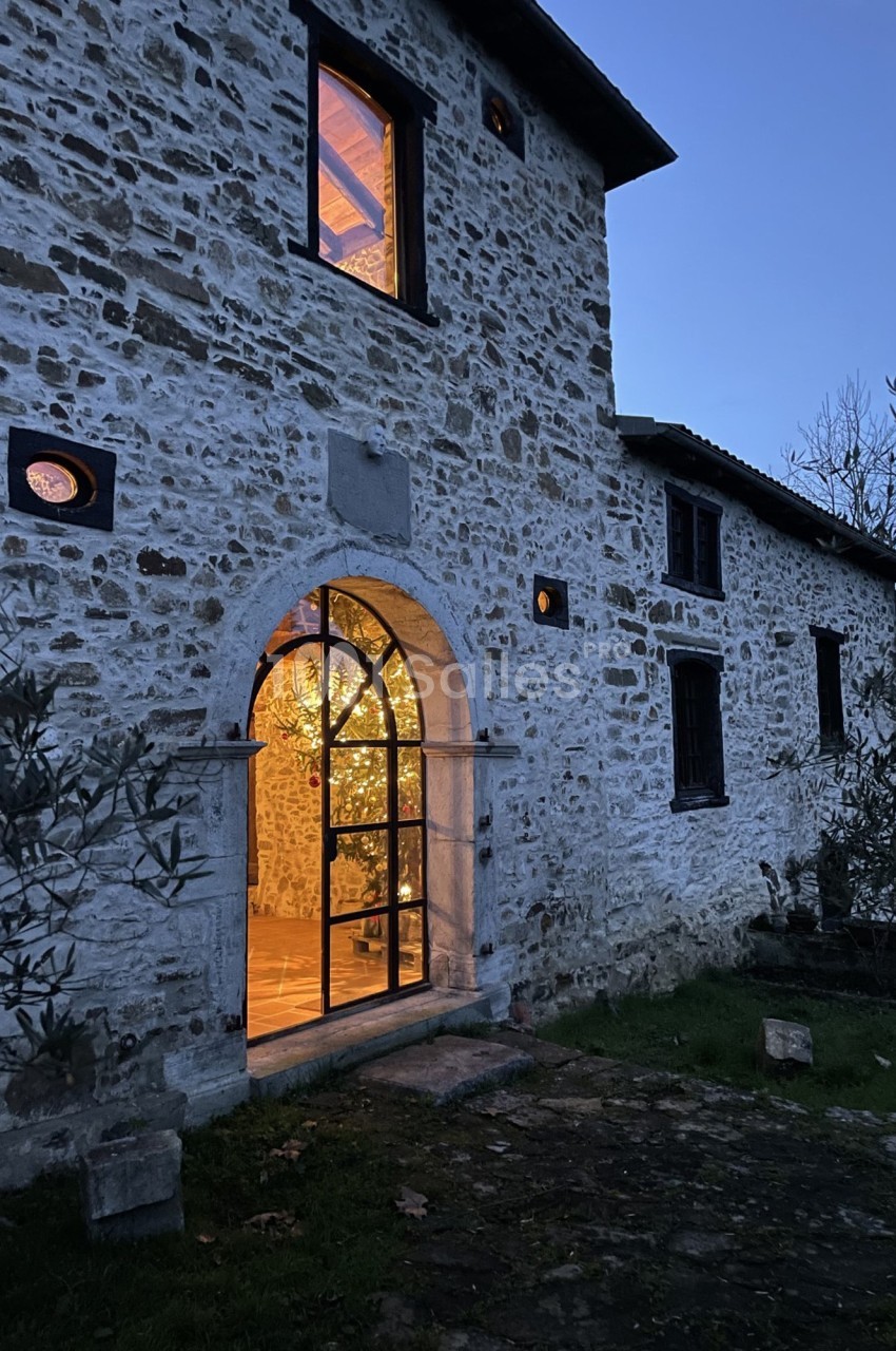 Façade en pierre d'une maison éclairée à la tombée de la nuit, avec une porte vitrée laissant entrevoir un intérieur…