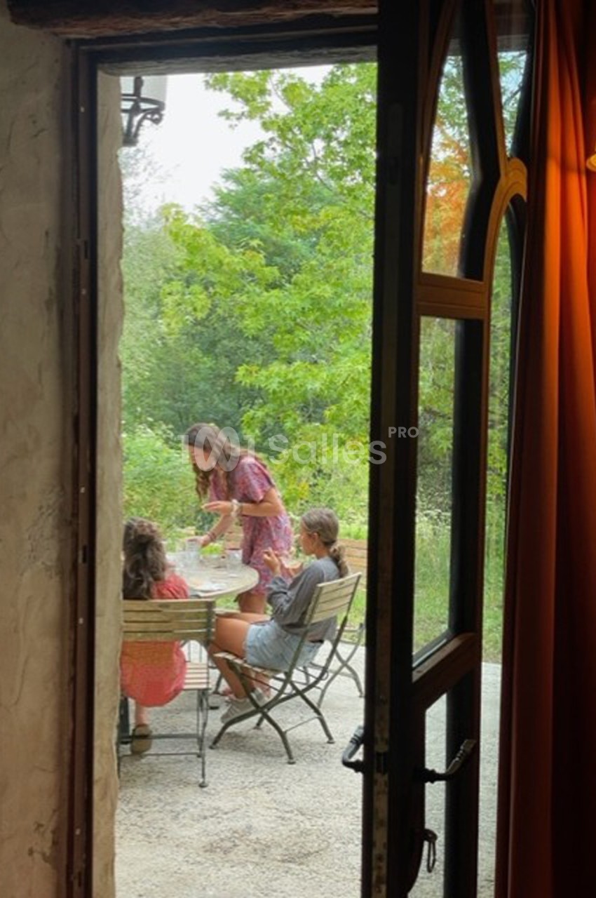Vue depuis une porte ouverte sur trois personnes assises à une table en extérieur, entourées de verdure.