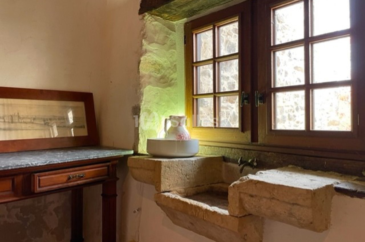 Évier en pierre ancienne sous une fenêtre à carreaux, avec un pichet blanc posé sur une vasque.
