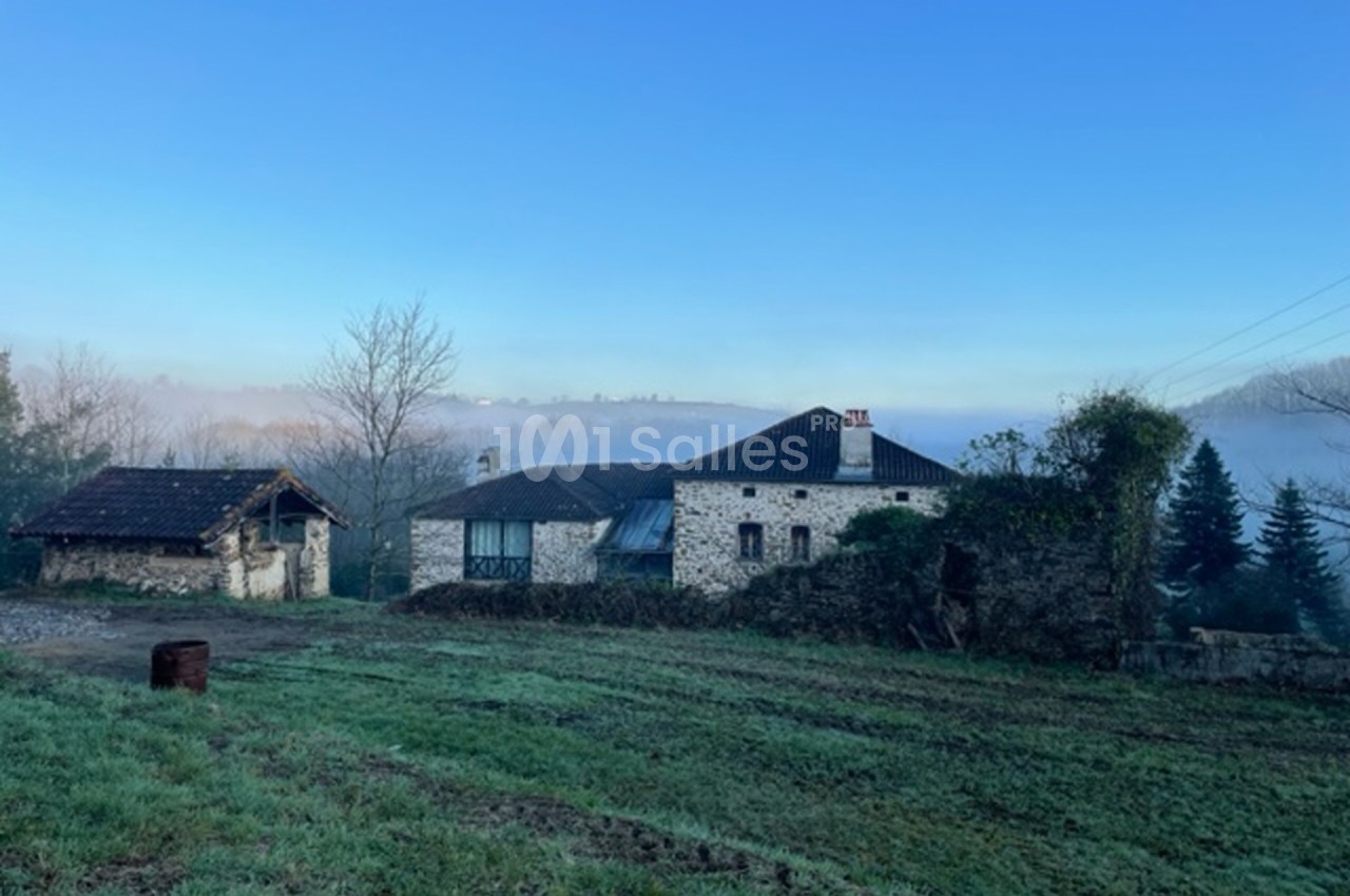 Maison en pierre entourée de verdure avec un ciel bleu dégagé et un paysage légèrement brumeux en arrière-plan.