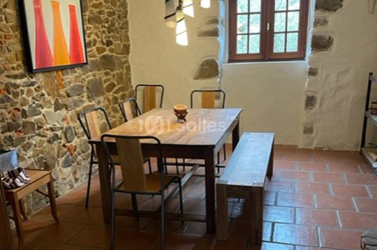 Salle à manger avec table en bois, chaises métalliques, banc, murs en pierre et fenêtre laissant entrer la lumière.